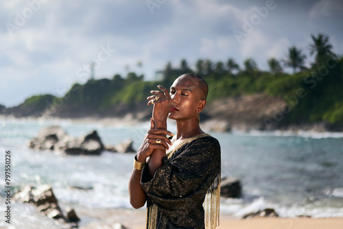 Epatage androgyne gay black man in luxury gown poses on scenic ocean beach. Non-binary ethnic fashion model in long posh dress and accessories stands with closed eyes in elegant posture.