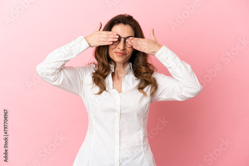 Middle aged caucasian woman isolated on pink background covering eyes by hands and smiling