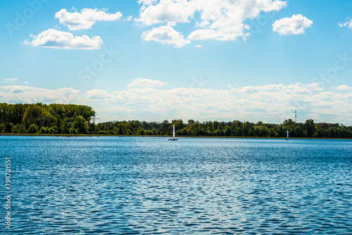 Ein Blauer Fluss an einem Waldstück mit weißen Wolken am blauen Himmel, ein Segelboot ist in der ferne zu erkennen