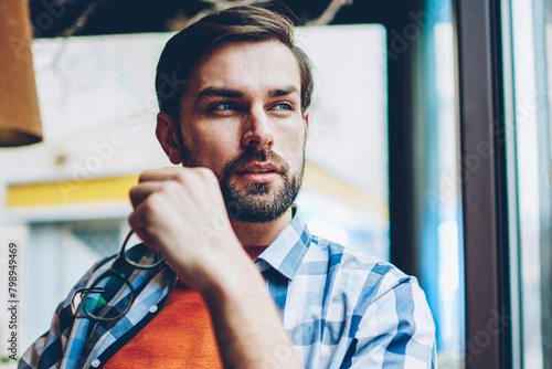 Dreamy handsome young man holding optical eyeglasses in hands while looking out of window and thinking on creative ideas for developing own startup project.Thoughtful hipster guy pondering on plans