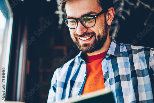 Concentrated successful hipster guy in eyewear smiling while reading excited plot of literature book in coworking space.Positive young man 20 years old enjoying favorite bestseller in free time