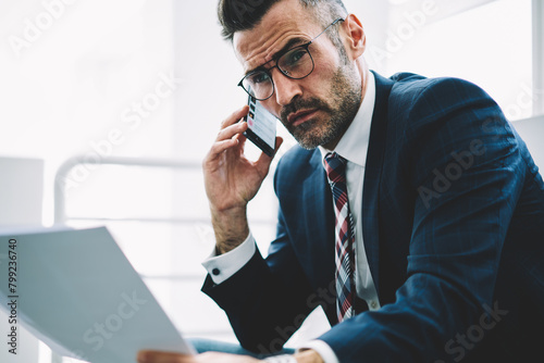 Confident successful businessman with financial text reports in hands calling to operator on smartphone working in office.Experienced proud ceo 50 years old with banking documents talking on telephone