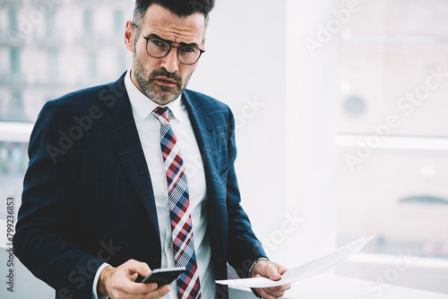 Half length portrait of confident mature proud ceo in suit looking at camera while dialing number on smartphone.Successful serious businessman with documents checking mail on cellular near copy space