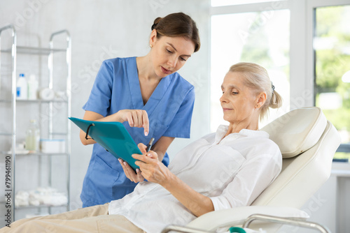 Young female doctor obtaining consent of patient for medical procedures or therapies. Focused elderly woman reading and signing medical agreement while sitting on examination couch..