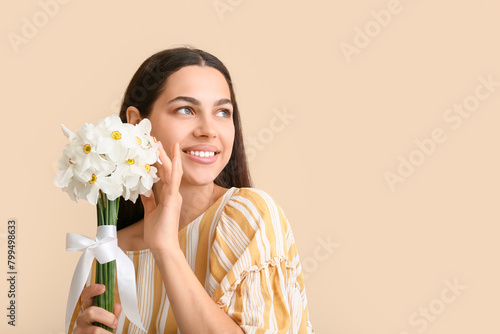 Beautiful young woman with bouquet of white daffodil flowers on beige background