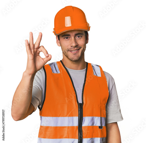 Young Hispanic worker in safety gear cheerful and confident showing ok gesture.