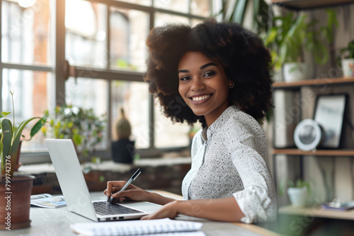 Happy african american woman with afro hair working on laptop writing notes in notebook at office desk, smiling positive female entrepreneur concept.