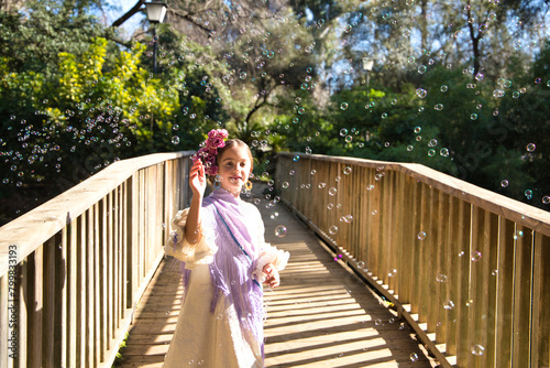A pretty girl dancing flamenco in a typical gypsy dress with frills and fringes walks on a wooden bridge in a famous park in Seville, Spain. The girl plays with soap bubbles.