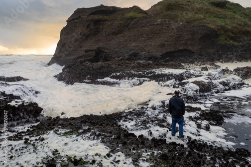 Man standing on rocks watching ocean waves and seafoam crashing over rocks at Piha, Auckland, New Zealand.