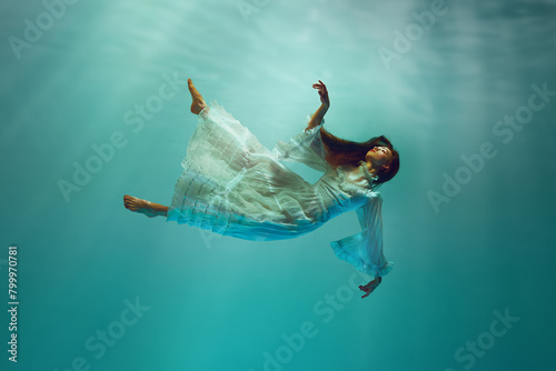 Finding inner peace. Serene underwater moment with elegant young girl in tender white dress levitating with calm expression underwater. Concept of surrealism, beauty, mystery and fantasy, freedom