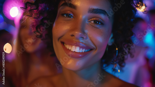 A young woman with curly hair is smiling at the camera. She is wearing a black dress and has a lot of makeup on. The background is blurred and there are colorful lights in the background.