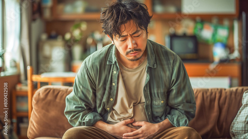  Asian man clutching his stomach have stomachache sitting on sofa , diarrhea problem concept