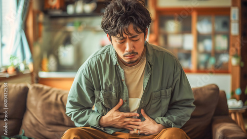  Asian man clutching his stomach have stomachache sitting on sofa , diarrhea problem concept