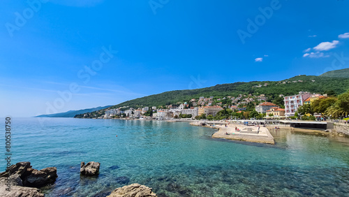 A panoramic view of the shore along Opatija, Croatia. There is a town located on the shore of the Mediterranean Sea. Calm surface of the sea. Stony beach. Green hills in the back. Sunny day. Cloudless