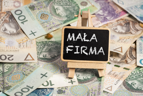  Black writing board on a wooden frame with the inscription "Mała Firma", Polish zloty PLN banknotes scattered in the background (selective focus) translation: small company