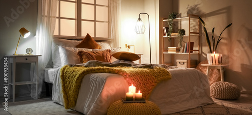 Interior of bedroom with cozy blankets on bed, burning candles and glowing lamp late in evening