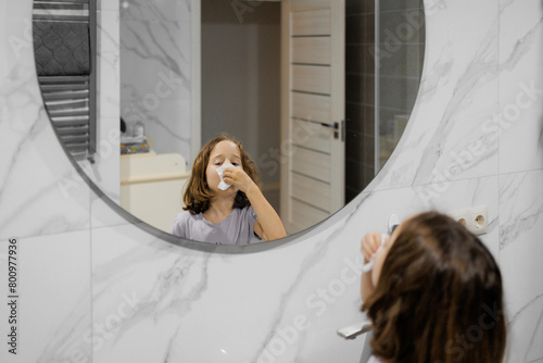 A young girl blows snot from her nose in front of a mirror