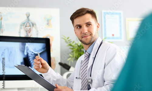 Portrait of smiling doc writing information on paper. Surgeon wearing medical uniform and stethoscope. X-ray picture of body on display of computer. Medical treatment concept. Blurred background