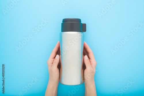 Young woman hands holding and showing closed new silver steel thermos with dark black plastic mug for hot drink or soup on pastel blue table background. Closeup. Point of view shot. Top down view.
