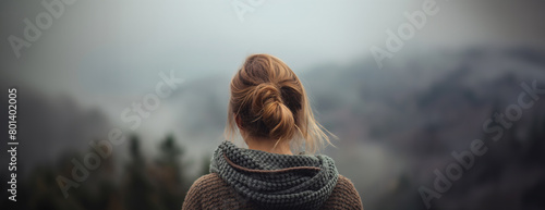 A woman with a bun hairdo is seen from behind, contemplating a foggy mountain range