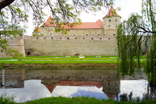 View of the Fagaras fortress in Romania: a landscape with a water-filled moat, trees, a defensive wall and a palace building behind it.