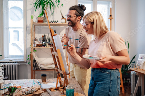 Affectionate smiling artistic couple painting on easel at atelier.