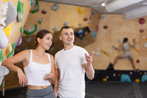 Two visitors of climbing hall are standing and relaxing, discussing active hobby. Guy and girl friends, standing and resting after overcoming obstacle course on artificial wall