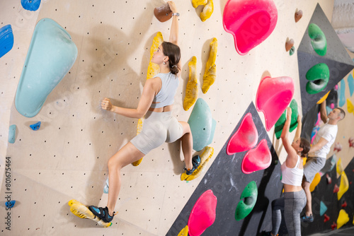 Young woman climbs steep artificial wall in sports complex and trains endurance and muscle strength. Active recreation in fitness center gym, modern types of recreation