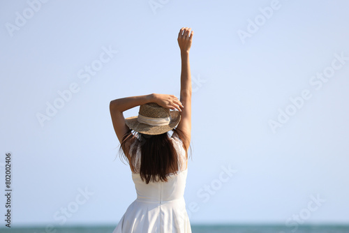 Young attractive ethnic Thai millennial woman posing at suny spreading arms to sides and various postures dressed in white casual clothes stands on the ocean with sandy beach. Love for travel