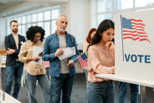 Asian woman standing in voting booth, voting, group of multinational US citizens waiting in line