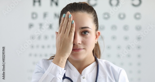Portrait of gorgeous woman ophthalmologist standing in clinic office and covering right eye with tender hand. Lady looking at camera with happiness. Eyesight check concept