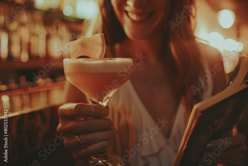 A smiling, fashionable woman is enjoying a crafted cocktail while deeply engaged in reading a book at a bar