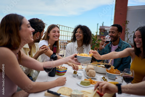 Group of multi-ethnic young friends enjoying meal gathered on rooftop. Happy millennial people having fun drinking red wine and eating celebrating a party event on summer day