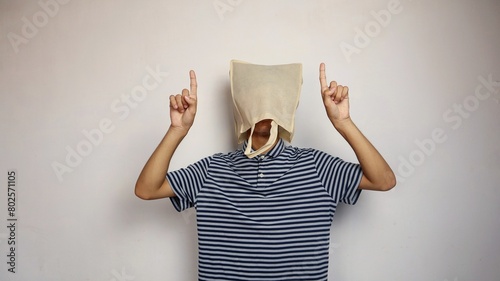 young Asian man wearing a shopping bag on his head with a hand gesture pointing upwards