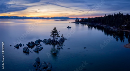 Lone tree on rocks at sunset on a calm Lake Tahoe. In the background os a forest and mountains covered in snow. Zephyr Cove, Nevada, United States of America.