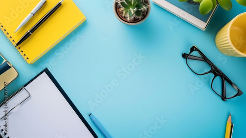 neat workspace with a yellow notebook, glasses, pens, a cactus, clipboard and a cup on a vibrant blue background.
