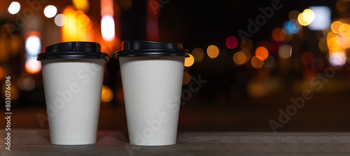 Close-up of two white eco-friendly recycled paper cups with coffee or tea standing on a bench against the background of a blurred night city with lights. Take the coffee with you. Banner