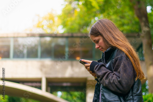 mujer joven con mirando su celular en la naturaleza. chica enviando mensajes con su celular con bosque de fondo 