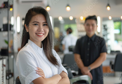 A smiling Thai smart woman hairdresser standing in her salon, looking at the camera with a relaxed and confident smile. In front of her is an empty chair for new clients to come sit in