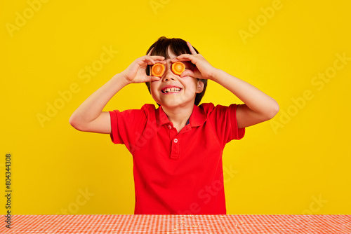 Cheerful little boy in red polo shirt using carrot slices as pretend glasses against yellow background. Creative fun with food. Concept of food, childhood, emotions, meal, menu, pop art