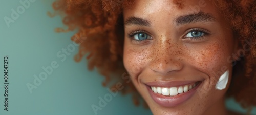 Within the beauty paradigm, a delighted black woman sporting red hair and freckles radiates happiness against a green wall, her face turned to the side, with a white cream noticeable on her complexion
