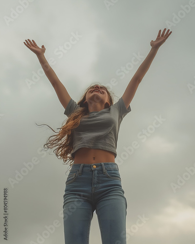 Photo d'une jeune femme en jean levant les bras, ciel gris en arrière plan, parfaite illustration de la liberté et de la jeunesse