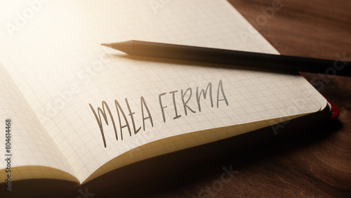 A handwritten inscription "Mała firma" on a grille of an open notebook on a wooden countertop, next to a black pencil, lighting of light. (selective focus), translation: Small company