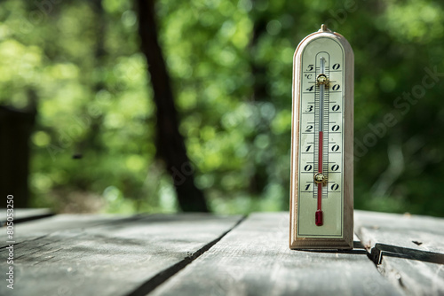 Termometr wskazujący wysoką temperaturę ustawiony na drewnianym stole na tle zieleni lasu.