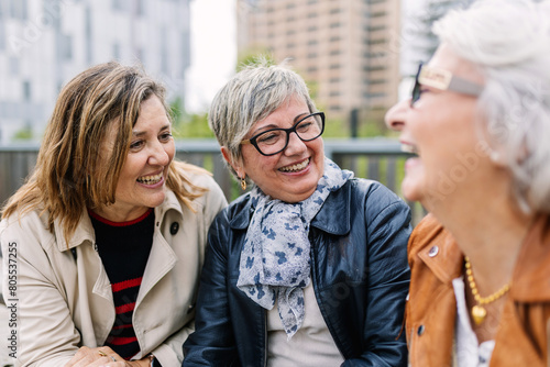 Three mature retired women laughing while talking sitting outside. Small group of mature females having fun enjoying a conversation bonding at city street. Elderly friendship concept.