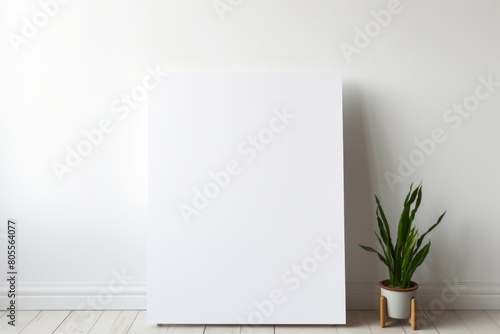 A blank white canvas positioned against a plain white wall in a room, with a potted green plant to the right.