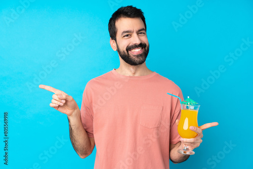 Young man over holding a cocktail over isolated blue background pointing finger to the laterals and happy