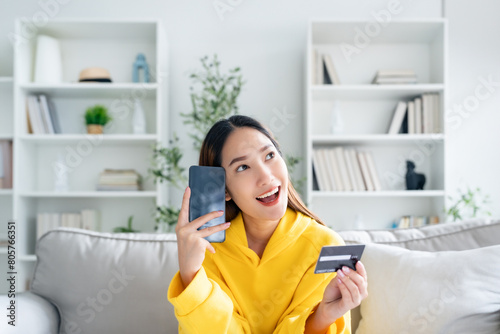 Young asian woman lying on sofa in living room, makes online banking payments through the internet from bank card on cell phone. Shopping online on mobile with credit card