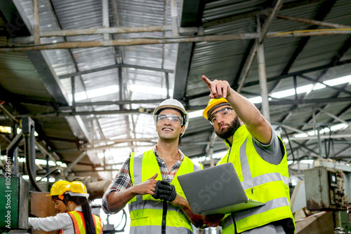 Industrial foreman and worker with helmet and safety vest using laptop computer during discussing together at manufacturing industry factory. Engineer inspecting workplace and checking process.