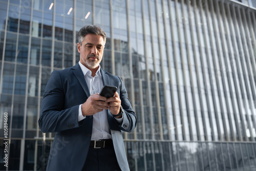 Mature hispanic senior business man using smartphone cellphone at office building. Focused serious latin middle age male entrepreneur businessman holding mobile cell phone for trading. Copy space